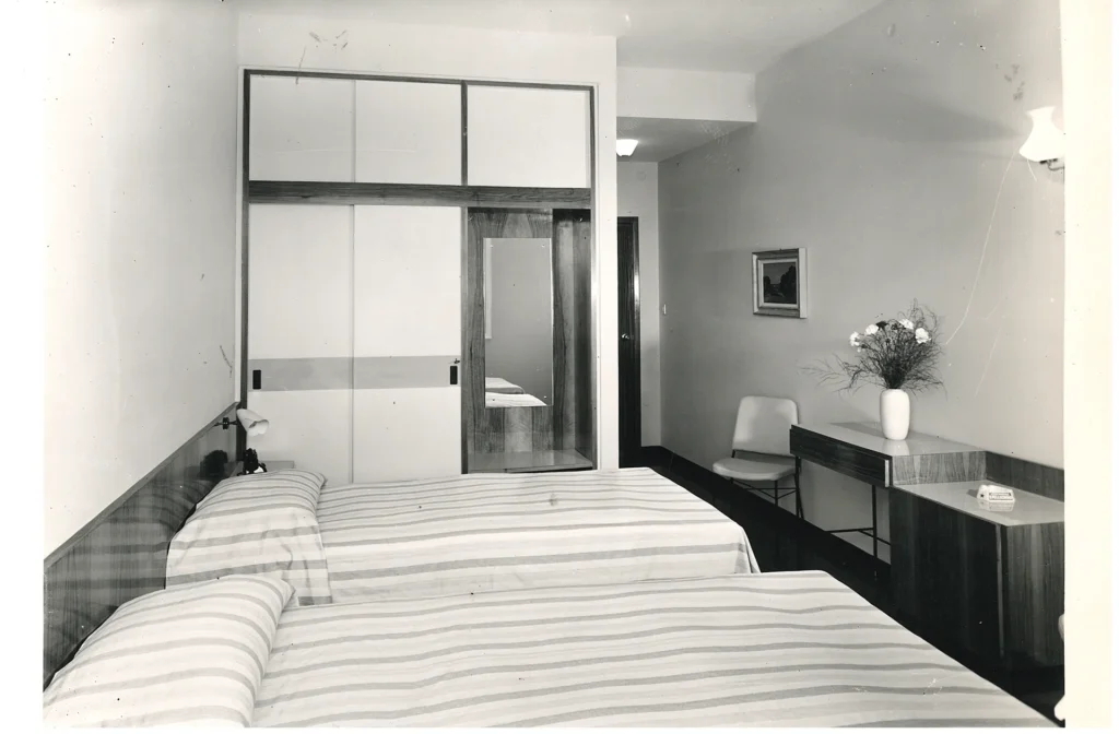 Nell'immagine una foto d'epoca delle camere dell'hotel. Al centro ci sono due letti con copriletti a righe. Di fronte c'è una scrivania con un vaso di fiori, un armadietto basso e una sedia. La parete accanto ai letti è occupata da un armadio a muro con specchio e accanto c'è la porta d'ingresso. La foto è in bianco e nero