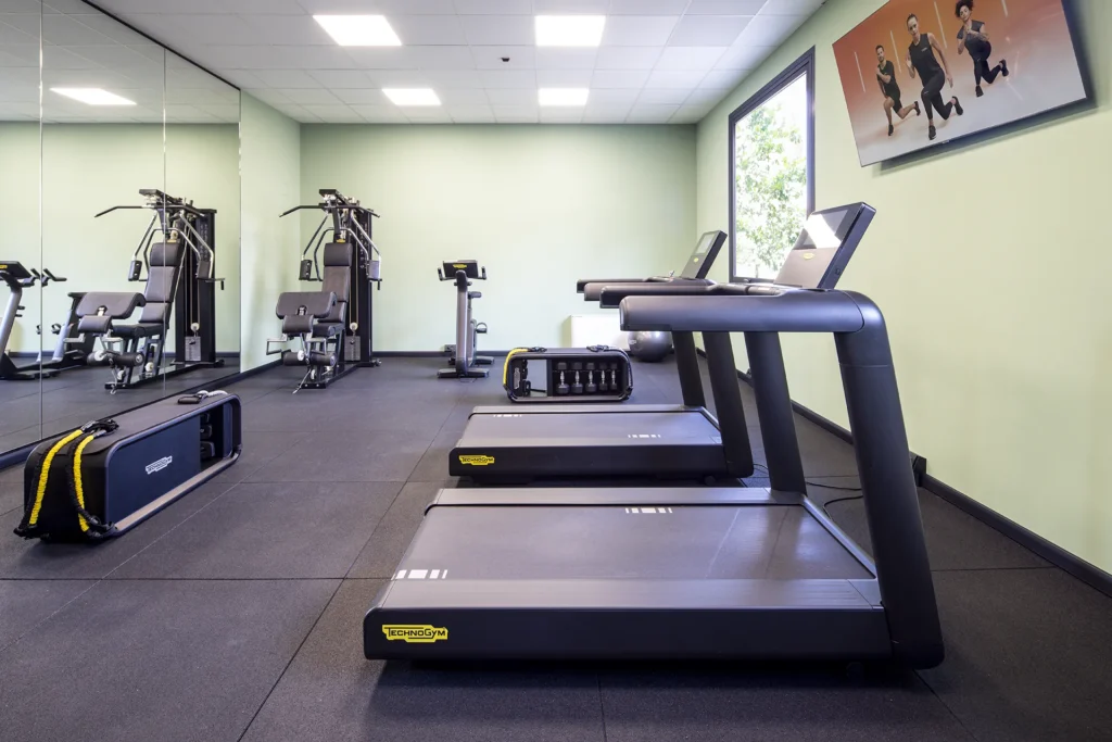 The image shows the hotel gym with various equipment and machines. In the foreground are two treadmills. To the left and behind the treadmills are two rectangular black containers for weights. At the back of the room is an exercise bike and a multifunctional machine.