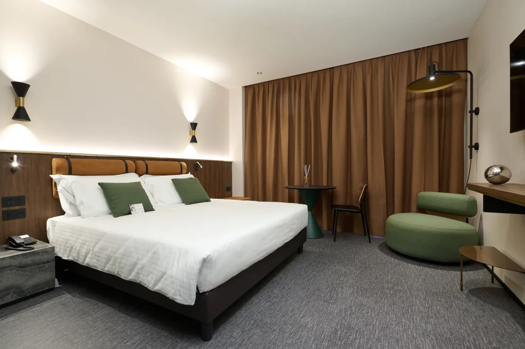 Nell'immagine la camera deluxe dell'hotel. A sinistra c'è il grande letto king size con doppi guanciali e cuscini decorativi verdi. Su entrambi i lati c'è un comodino con luce da lettura. Dietro al letto c'è un pannello decorativo di legno fino a metà altezza e la testata è composta da due cuscinoni arancioni. Sulla parete sopra al letto ci sono due appliques. Di fronte al letto c'è una mensola e accanto una poltroncina verde tondo. Sopra la poltroncina appesa al muro c'è una lampada nera. In fono alla stanza c'è un tavolino tondo nero e verde con sedie e dietro di essi delle tende marroni chiuse