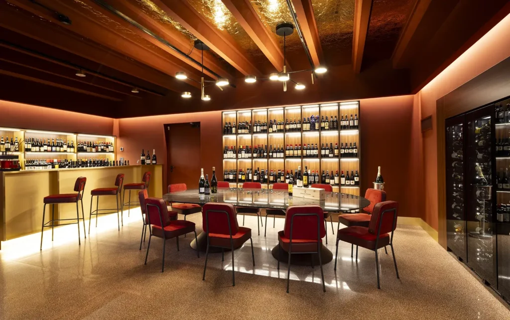 The image shows the restaurant's party & fashion wine room. In the center is a large elliptical table surrounded by red velvet chairs. To the right is a wine cellar and a wall-mounted wine display. To the left is the bar counter with three red velvet stools. Beyond the counter are illuminated shelves lined with wine bottles. The back wall also features a brightly lit shelving unit with wine bottles.