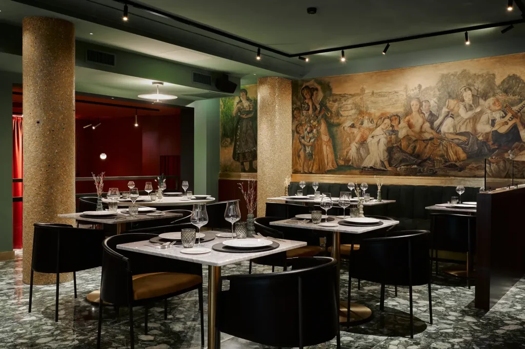 Nell'immagine il ristorante dell'hotel. Ci sono dei tavolini apparecchiati per la cena, sedie di pelle nera e sedile giallo e una lunga panca in fondo al centro. Le pareti sono affrescate