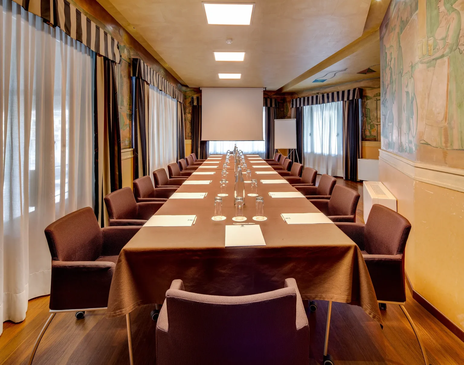 Nell'immagine la sala meeting Pendini dell'hotel. La sala è occupata da un grande tavolo rettangolare con tovaglia marrone e sedie di velluto marrone intorno. Sul tavolo ci sono delle cartelline, bicchieri e bottiglie d'acqua. Sui lati ci sono delle finestre coperte da tende e le pareti sono affrescate. In fondo alla sala c'è un telo per proiezione. Il pavimento è di legno scuro.
