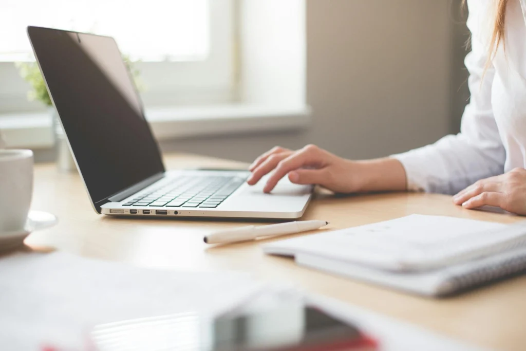The image shows a woman's hands working on a computer keyboard on a wooden table. Next to the computer is a notepad, a pen, and a cup.
