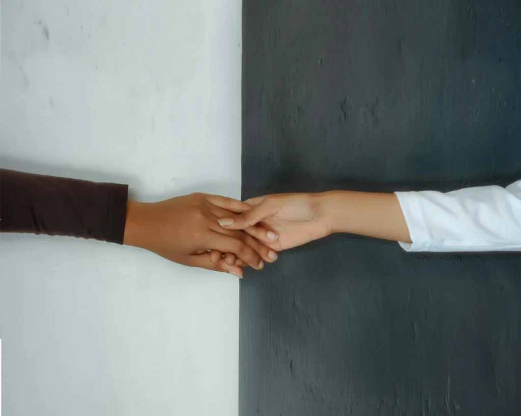 The image shows two hands clasping. The wall is split in half at the point where the hands are clasped: the right is black, the left is white.