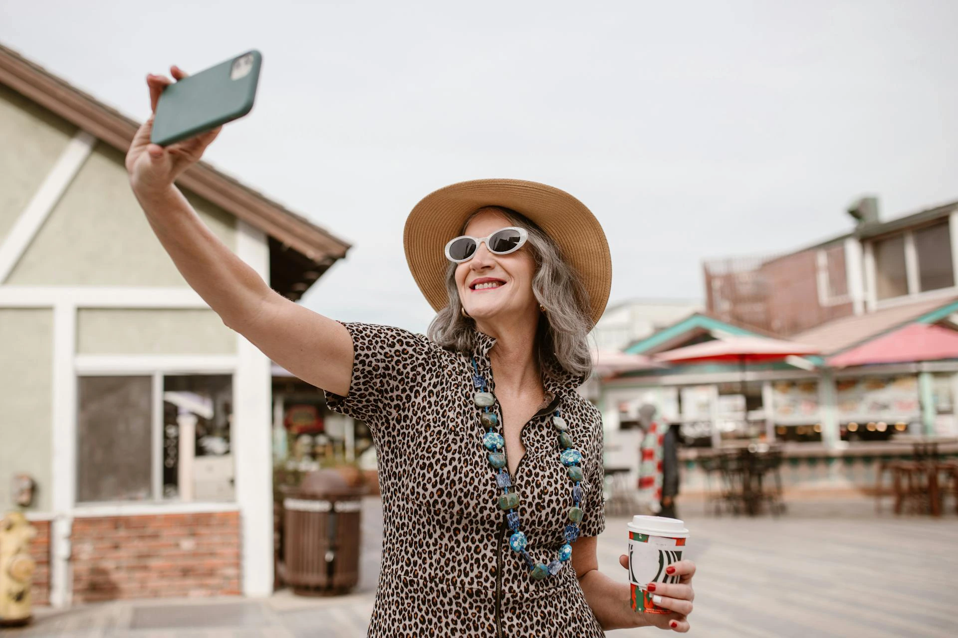 The image shows a woman smiling and taking a selfie. She's wearing an animal-print zip-up shirt, white sunglasses, a straw hat, and a necklace with large blue stones. She's holding a takeaway coffee.