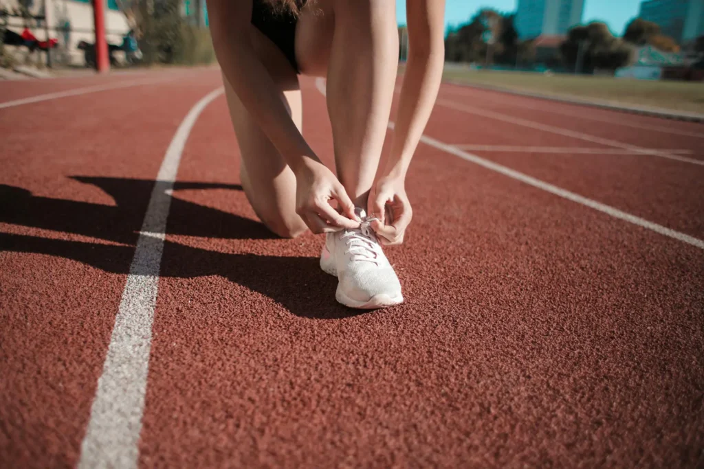 The image shows a girl tying her white running shoes. She stands on an orange running track.